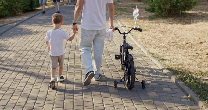 Father walks on paved path holding hand of son while bicycle rides nearby. Boy steps with confidence while father leads steady walk through park holding item at sunset