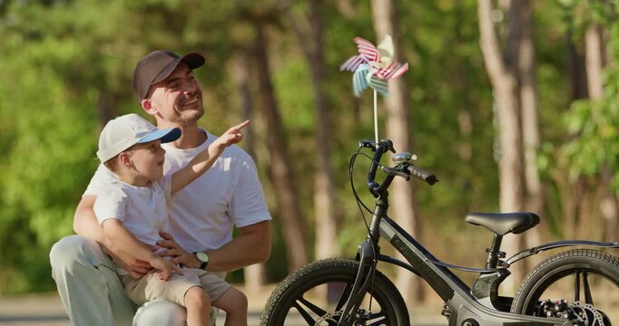 Father relaxes beside bicycle laughing loudly during cheerful pause with son. Boy leans toward parent smiling widely as father amuses with gestures in forest park