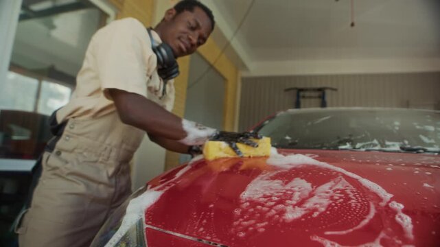 Tilt up of African American mechanic in work overalls and gloves using sponge with soap to wash hood of red car inside garage