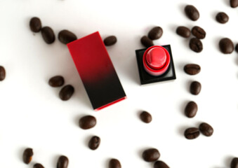 Red lipstick in open case surrounded by scattered coffee beans on white background, stylish composition symbolizing beauty, elegance and lifestyle.