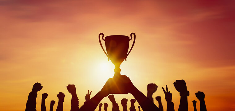 Silhouette of people's hands celebrating victory and success with a trophy on sunset background. Concept of joint celebration of victory and teamwork achieved the organization's highest goals.