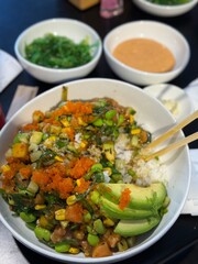 Bowl filled with sushi rice, topped with avocado, edamame, corn, and fish roe, surrounded by dipping sauces and green onions, highlighting delicious food presentation