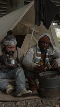 Vertical shot of Black male vagabond setting fire to cardboard box in small steel barrel and warming hands over it with Caucasian friend in homeless camp