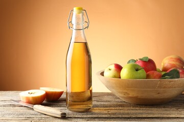 Delicious cider in glass bottle, apples and knife on wooden table against orange gradient background