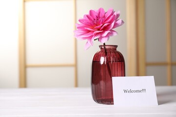 Card with word Welcome and vase with flower on white wooden table indoors. Space for text
