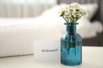 Card with word Welcome, vase with flowers and towel on white table indoors