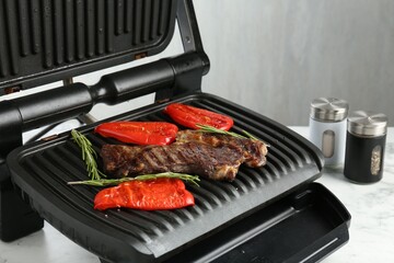 Electric grill with beef steaks, bell peppers and rosemary on white marble table, closeup