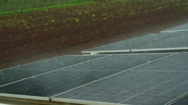 A focused shot of rain bouncing off rows of solar panels in a farmland, with wet reflective surfaces and green fields in the background in Lucban, Quezon Province Philippines