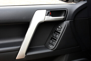 Close-up of modern car interior door with metallic handle, black textured panel and integrated power window control buttons, showing sleek automotive design and functionality.