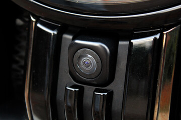 Close-up of black car front camera integrated into glossy grille, showing round lens, modern automotive technology and sleek design detail of vehicle safety system.