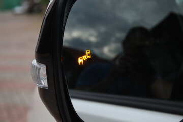 Car side mirror with illuminated orange blind spot monitoring warning indicator, glossy black frame and clear turn signal light on modern vehicle exterior.