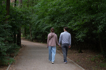 A couple enjoys a peaceful stroll down a shaded path in a vibrant summer park. Seen from behind, they are immersed in conversation amidst the lush green foliage and dappled sunlight. The image evokes 