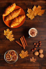Top-down view of a cozy autumn breakfast setup featuring croissants, a mug of cinnamon coffee, dried oranges, nuts, cinnamon sticks, and maple leaves on a wooden table.