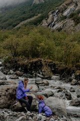 Fototapeta premium Young Caucasian man and a child sit on rocks by a river in a mountainous area. Both wear purple jackets. Lush greenery surrounds the scene.