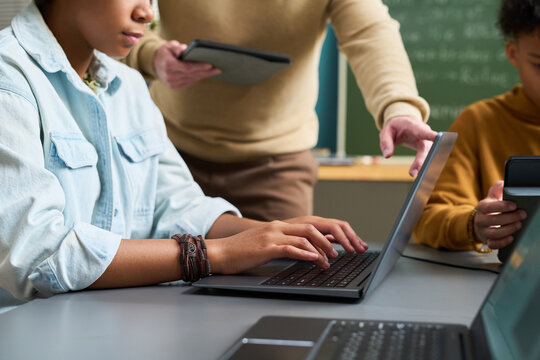 Teenage Black girl typing on laptop while Caucasian man holding tablet instructing and another teenage Black girl using digital tablet in classroom setting