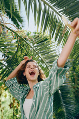 Joyful woman stretches arms and laughs under lush palm leaves, a bright outdoor moment captured in warm sunlight.