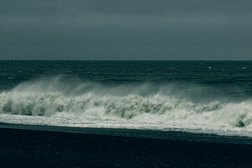 The image captures a dramatic scene of crashing waves upon a dark, rocky shore under a moody sky, conveying a sense of power and nature's intensity.