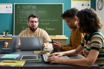 Caucasian young adult man explaining AI prompting to two multiethnic teenagers using laptops in...