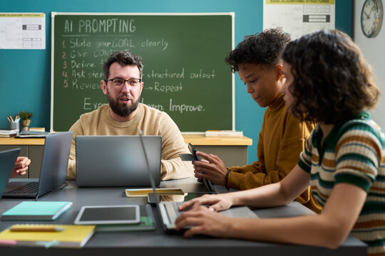 Caucasian young adult man explaining AI prompting to two multiethnic teenagers using laptops in classroom, students listening attentively and taking notes with digital devices