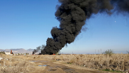 A large plume of black smoke rises from a fire in a junkyard or waste site. The image highlights...