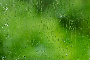 Water drops run down the glass surface with rain. An atmospheric rain picture. Abstract wallpaper. Summer greenery behind the glass. Close-up of the drops.