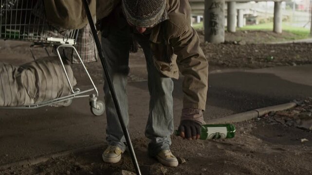 Tilt up view of Caucasian male vagabond with walking stick picking up empty glass bottle from ground, examining it, putting in shopping cart and pushing it