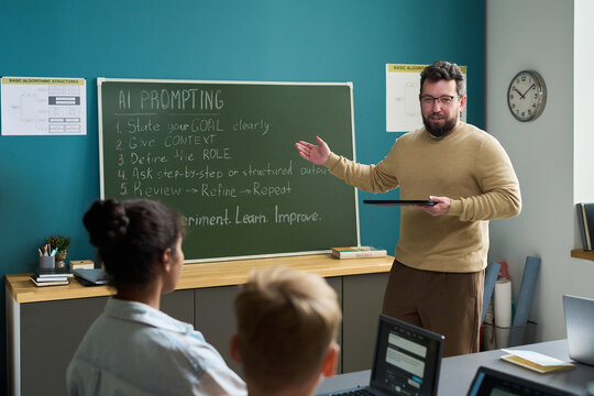 Caucasian man standing at chalkboard holding digital tablet, explaining AI prompting steps to multiethnic group of teenagers using laptops in classroom setting