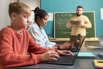 Caucasian teenage boy and Black teenage girl using laptops in classroom while male teacher standing near chalkboard holding digital tablet in background