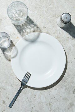 empty white plate on a grey terrazzo background with transparent glasses, fork and salt shaker
