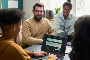 Obraz premium Caucasian man and two Black teenagers sitting at table collaborating on laptops, smiling and engaging in group discussion during teamwork session in modern office
