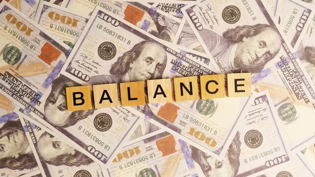 Wooden blocks with the letters "Balance" arranged on a slowly rotating stack of dollar bills. Looping 360-degree of wooden blocks with the words "Balance" as a business financial concept. 4K footage.