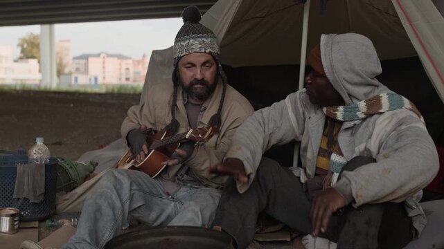 Tilt up view of two multiethnic male hobos relaxing near fire barrel and tent under bridge, Caucasian man playing ukulele, his African American friend bopping head to beat