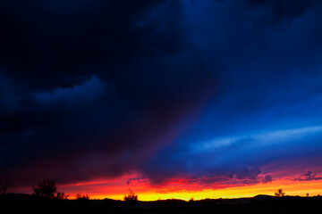 A breathtaking sunset after rainfall in Mongolia, where dramatic clouds glow in fiery red and deep blue hues. The sun sets like burning fire behind the horizon, casting mountains and trees in striking