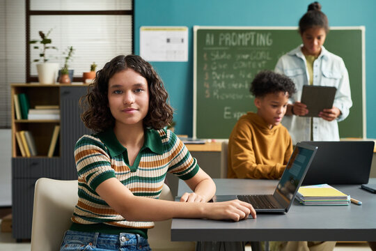 Portrait of Caucasian teenage girl sitting at desk using laptop while looking at camera, multiethnic teenagers in background working together with digital tablet and books - Powered by Adobe