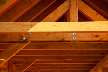Interior view of a wooden roof structure with visible beams and bolts, illuminated by warm sunlight. The natural wood texture highlights craftsmanship and sustainable building.