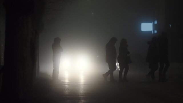 A group of people walking on road crossing in the dark night with fog and stopped car in the background. Scene is mysterious and eerie