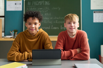 Two teenage boys, one Black and one Caucasian, sitting at classroom desk using digital tablet, both looking at camera with relaxed expressions, chalkboard visible in background