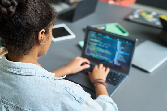 Black teenage girl sitting at desk typing on laptop computer with code visible on screen, working on programming project in modern classroom or collaborative workspace