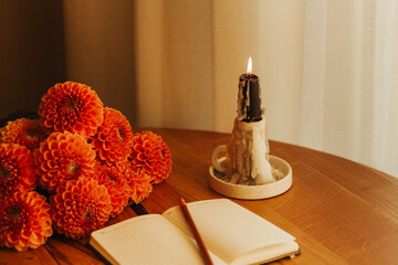 Cozy autumn still life with burning candle, notebook, pencil and orange dahlia flowers on wooden table. Warm home decor, fall mood, hygge atmosphere, relaxation and mindfulness concept.