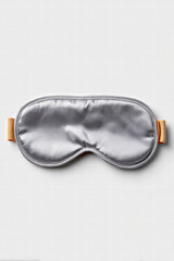 Silver satin sleep mask with orange elastic strap laid flat on white background, soft padded texture conveying calm relaxation and comfortable night rest