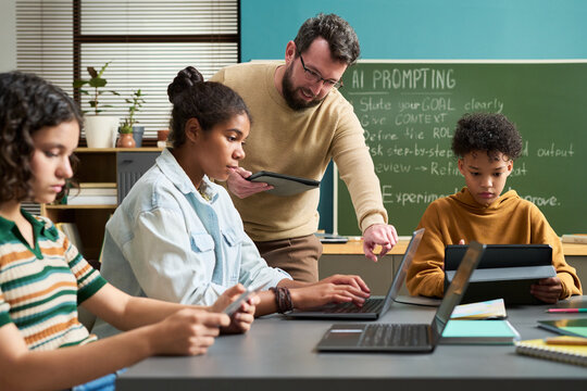Caucasian man instructing multiethnic teenagers using laptops and tablets in classroom, students focusing on digital devices while teacher providing guidance near chalkboard