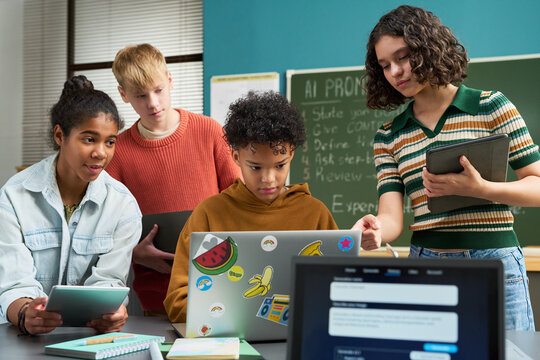 Group of multiethnic teenagers collaborating on digital project in classroom, using laptop and tablets, standing and sitting around desk, focused on technology and teamwork