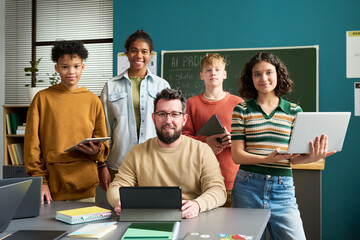 Group of diverse teenagers standing with Caucasian man holding digital tablets in classroom, teenagers smiling and looking at camera, teacher sitting at desk with laptop, chalkboard in background