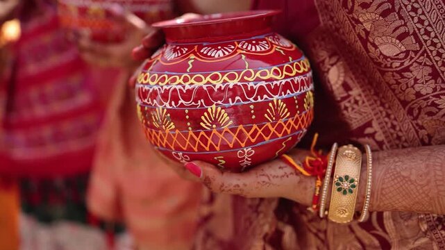 Close-up: Woman's henna hands in saree holding decorated red pot (Kalash/Matka) during traditional Indian wedding/puja ceremony.