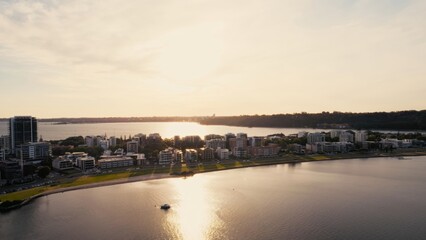 Obraz premium Waterfront city scene at sunset showcases tranquil atmosphere. A boat gently floats on calm waters reflecting the warm hues of the sky.