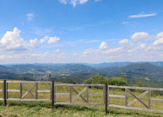 Fototapeta premium A vast mountain landscape, framed by a wooden fence, conveys the grandeur of nature. Green hills and forests, smoothly transitioning into the blue outlines of mountains on the horizon, create a sense 