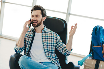 Smiling man talking on the phone while waiting at the airport terminal with a backpack and luggage for his flight