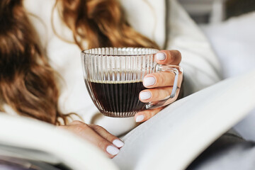 Young woman holding glass cup of coffee and reading book. Relaxed morning routine at home. Lifestyle detail scene. Cozy home atmosphere. Hot black drink in transparent elegant cup in female hand.