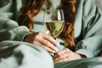 Relaxed woman holding glass of white wine. Casual home lifestyle detail. Green clothing cozy moment. Alcoholic drink concept. Hand with manicure and jewelry accessory holding elegant glassware.