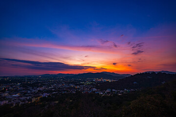 A scenic view of a town nestled among lush green mountains at sunset, with a temple structure perched on the hillside and a vibrant orange sky creating a warm, tranquil atmosphere.